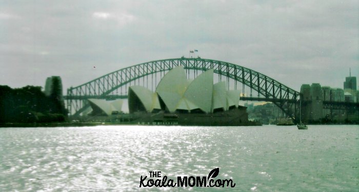 Sydney Opera House sits in front of the Sydney Harbour Bridge as sunlight sparkles on the ocean.