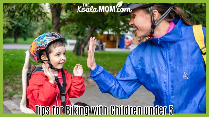 Tips for Biking with Children under 5. Mom in a helment and blue rain coat gives her son, also wearing a helmet and a red rain coat and sitting in a bike seat, a high five. Photo by Atlantic Ambience via Pexels.