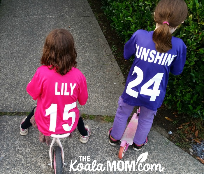 Girl on bike wearing a pink sports team jersey and girl on a scooter wearing a purple sports team jersey