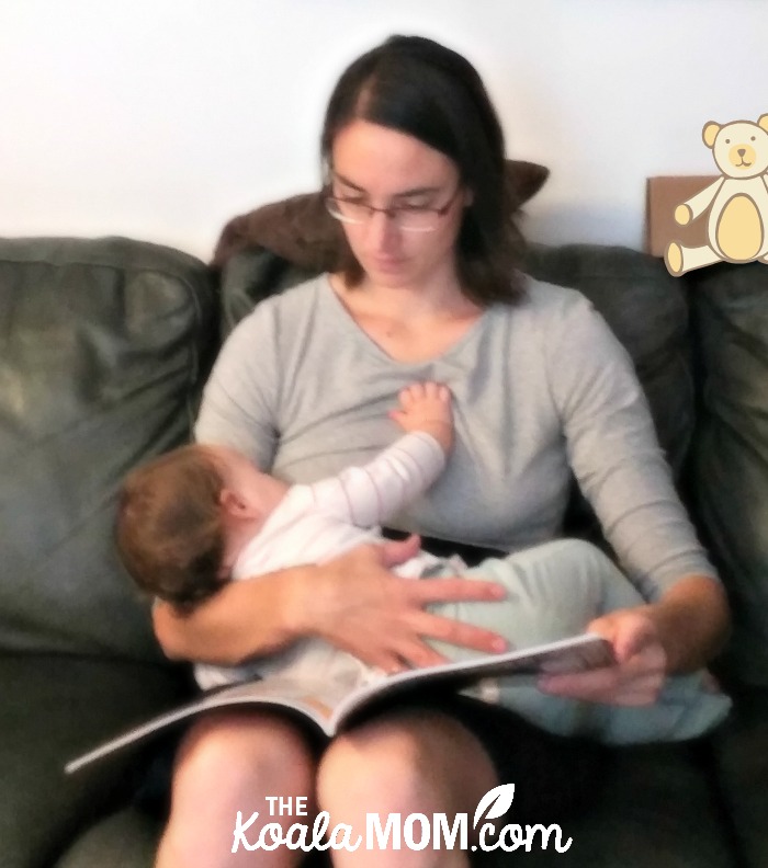 Mom wearing a blue-and-grey nursing dress from Momzelle while feeding a baby and reading a book