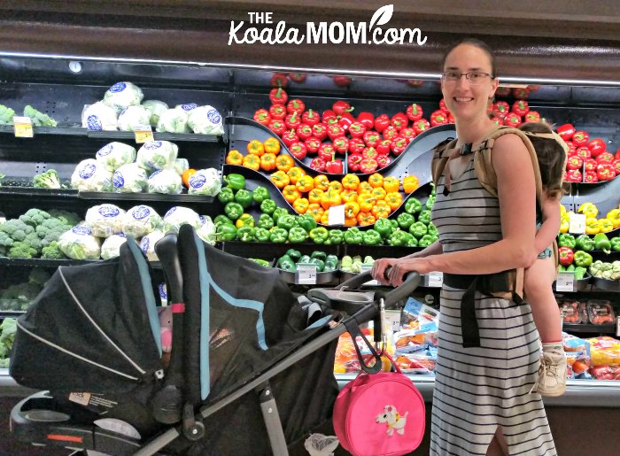 Mom at a grocery store with an infant in her stroller and a toddler in a baby carrier on her back.