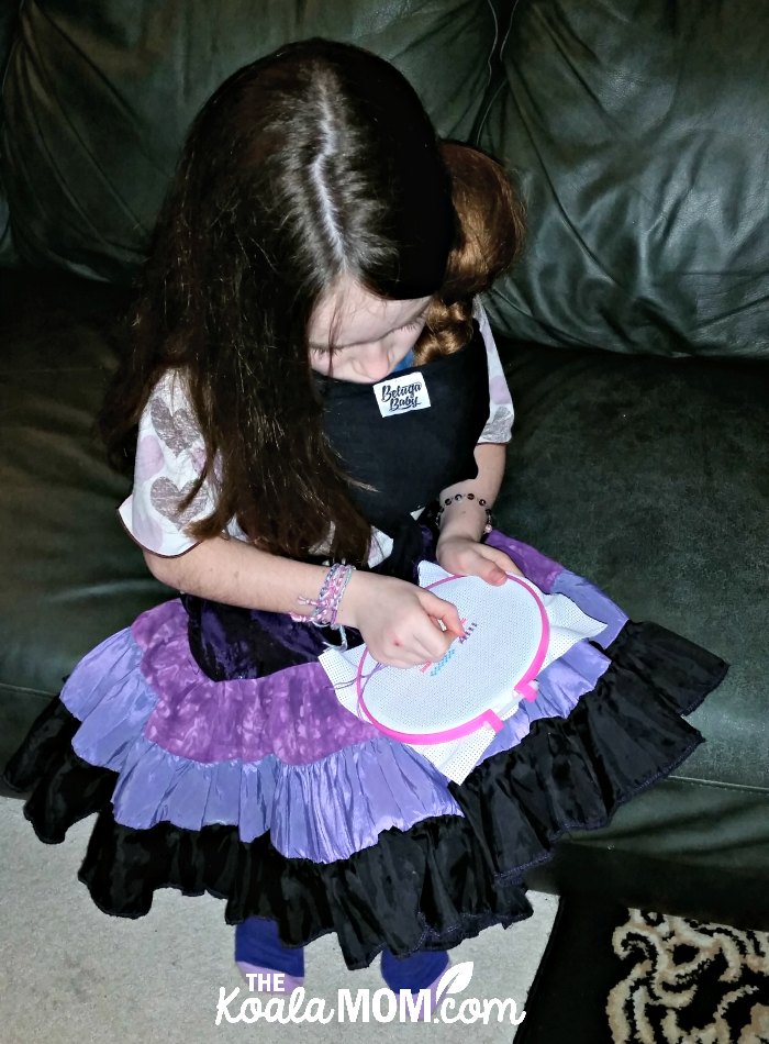 Nine-year-old girl wearing her 18-inch doll in a Beluga Baby bamboo doll wrap while doing cross-stitch