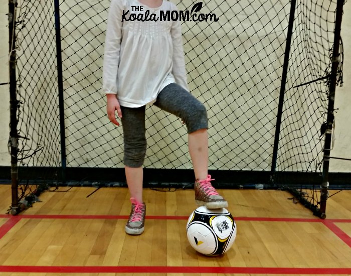 Girl standing in a soccer net with her foot on a Sportball soccer ball during one of their kids sports classes.