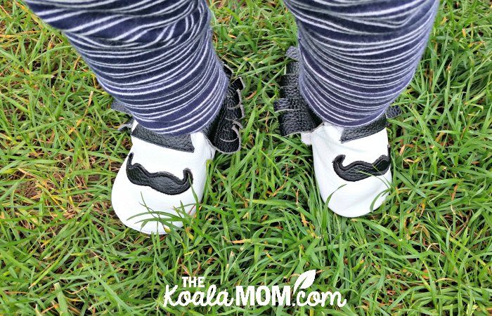 White baby moccasins with black mustaches on them; baby in blue-and-white pants standing on grass