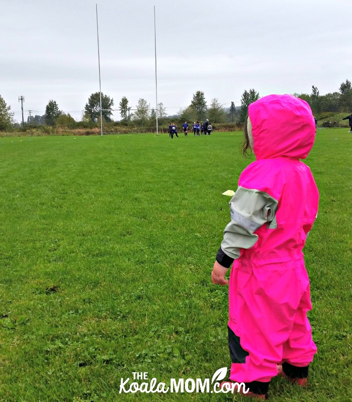 Toddler wearing her bright pink Oakiwear rain suit at her sisters' mini rugby game; the right gear can help encourage your kids to play sports.