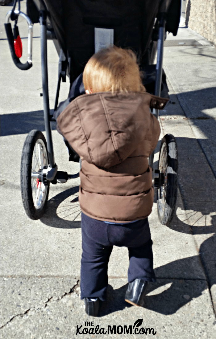 Baby pushing his stroller while wearing a brown winter coat on a walk with Mommy.