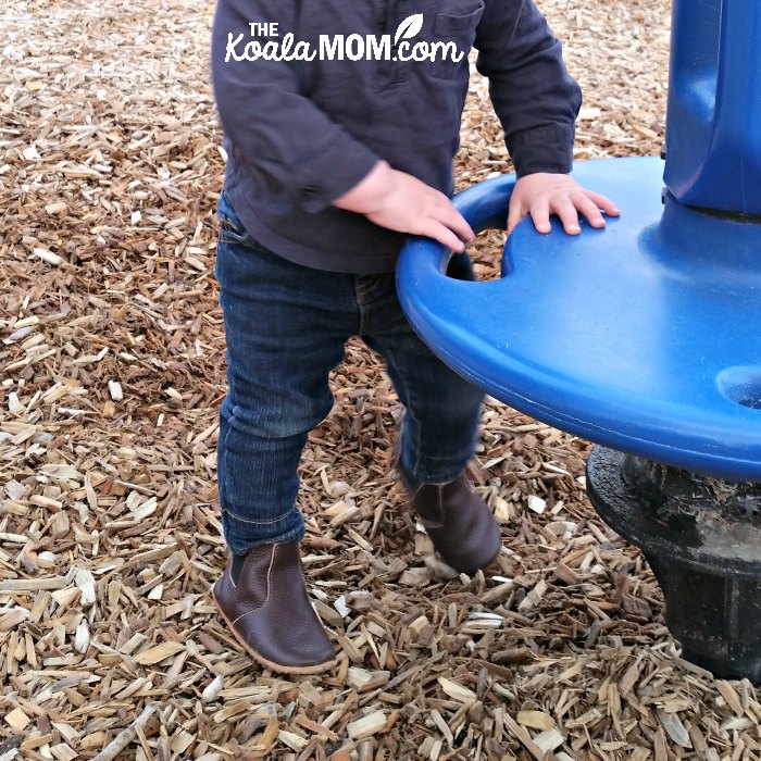 Baby standing beside spinning ride in his leather boot moccasins.