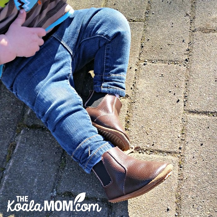 Baby wearing blue jeans and brown leather boots while sitting on cobblestones.