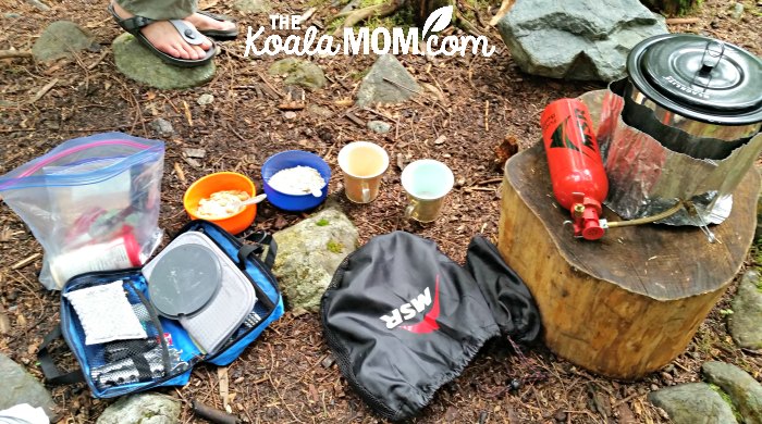 A backpacking kitchen on an overnight backpacking trip in BC: lightweight stove, fuel bottle, pot, utensils, and bowls with instant oatmeal and cups with instant coffee, waiting for water to boil.