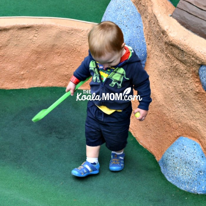 Toddler walking through mini golf course with a club and ball.