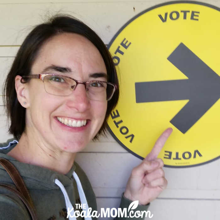 Smiling woman points to a yellow "VOTE" sign on a wall.