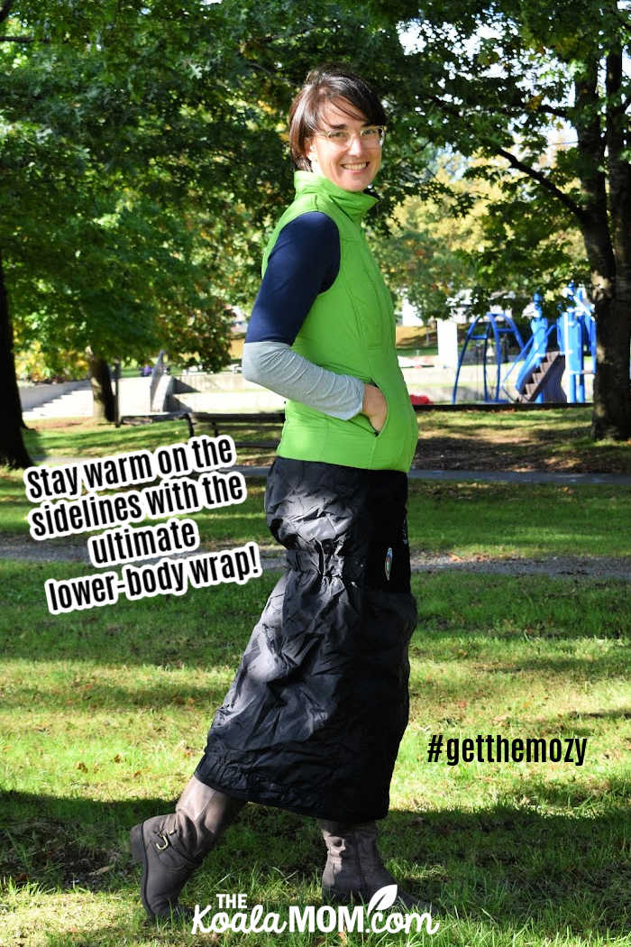 Stay warm on the sidelines with the ultimate lower body wrap! #getthemozy (Woman wears a green vest, boots, and a black Mozy - wearable blanket - wrapped around her waist.)