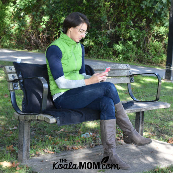 Woman sitting on the Mozy on a park bench while she reads a book on her phone.