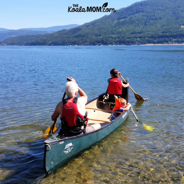 Toddler riding in a canoe with his big sister and grandma, while playing with a child-size paddle.