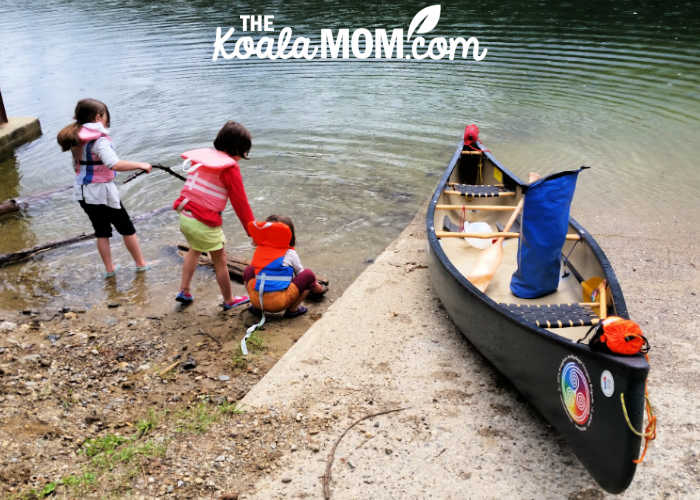 Three girls wearing their PFDs playing beside a canoe on a boat launch.