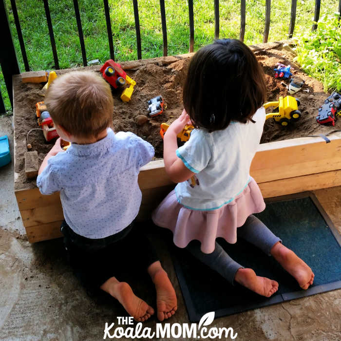 Two children playing with trucks together in a garden box.