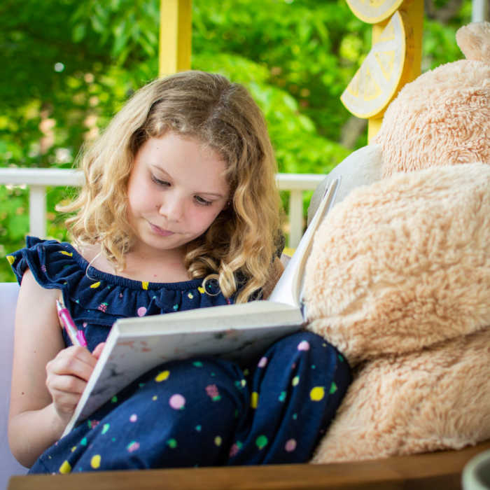 A young girl writes in her diary while sitting beside her giant stuffed teddy bear.