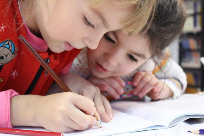 Two girls leaning over a notebook together, while one writes with a pencil.