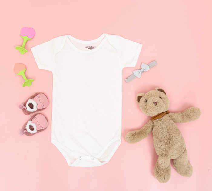 Flat lay of a baby onesie, shoes, teddy bear, and toys on a pink background.