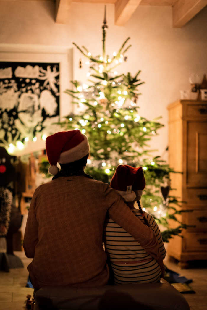Parent and child look at a lit Christmas tree while wearing Santa hats.