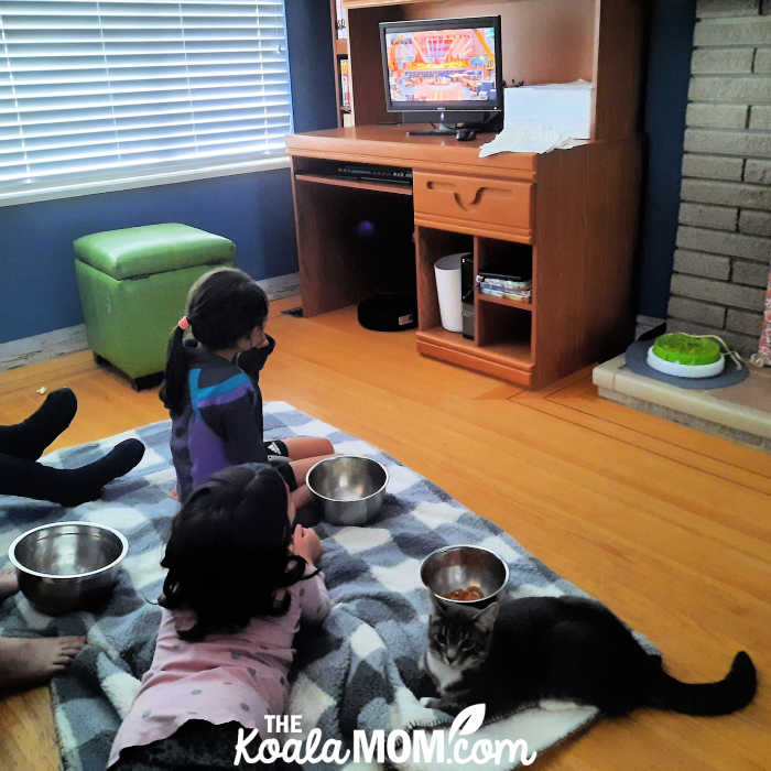 Two girls watching Sing 2 while sitting on a blanket eating popcorn with their cat.