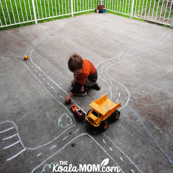 Four-year-old boy playing with his trucks on a sidewalk chalk road on his patio.