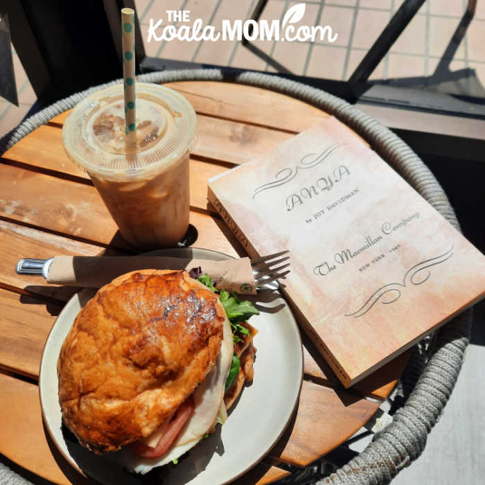 Anya by Joy Davidman, beside an iced coffee and sandwich on a sunlit table.