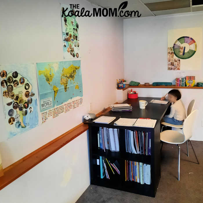 Homeschool student sits at a desk in the corner of her school room, with maps on the walls and a bookshelf nearby.