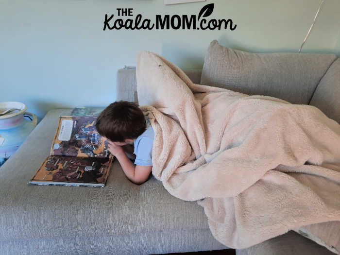 Child sprawls under a fuzzy blanket on a cosy sectional, reading a book. Photo by Bonnie Way.