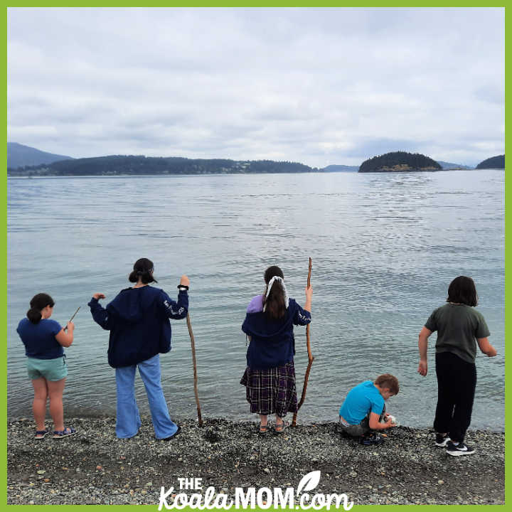 Five kids face the ocean, two holding walking sticks, one kneeling to look at a shell. Photo by Bonnie Way.