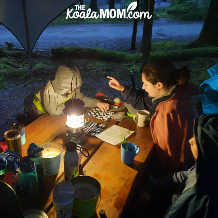 Kids playing Chess in the dark at a picnic table with the light of a lantern. Photo by Bonnie Way.