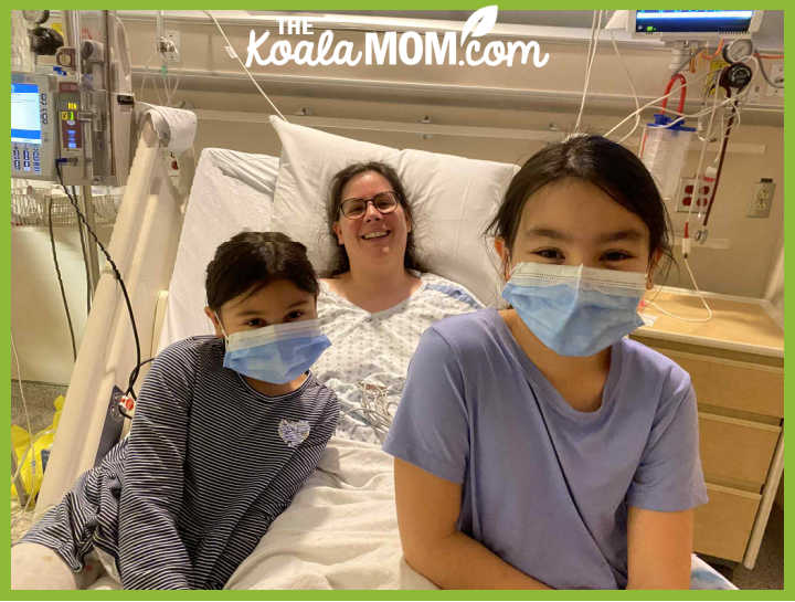 Photo of Shelby Emmanuels in a hospital gown and bed, with her masked daughters sitting with her. Photo provided by Shelby.