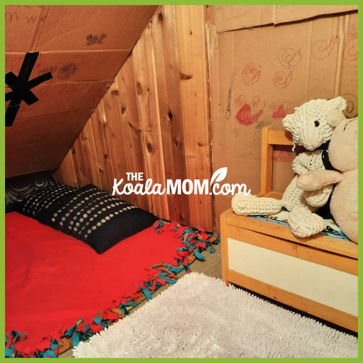 Kids play area under stairs with blanket, pillows, stuffed animals, and toy bench.