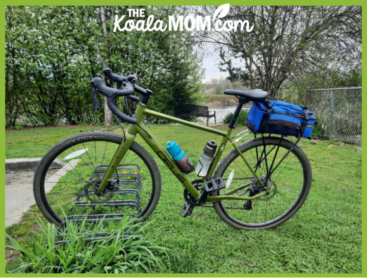 Photo of green gravel bike with blue rack bag and two water bottles sitting on grassy area by Bonnie Way.