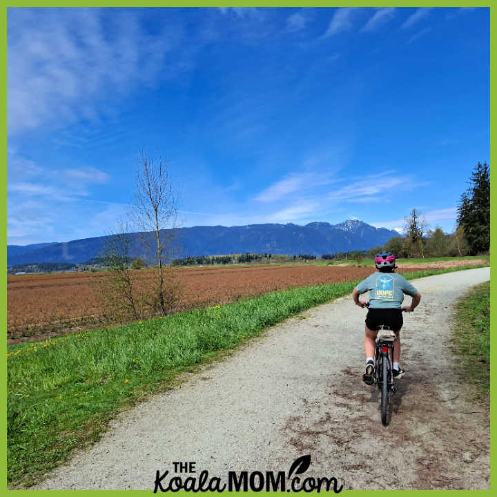 Photo of teenager cycling on gravel trail past blueberry fields with big blue sky above by Bonnie Way.