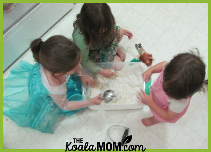 Photo of three girls sitting on flour, playing in a DIY flour sensory bin, by Bonnie Way.
