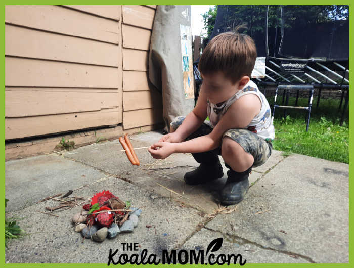 Boy holds two pepperoni sticks over an imaginary "campfire" made of his red stuffed lizard and some rocks, on a patio. Photo by Bonnie Way.