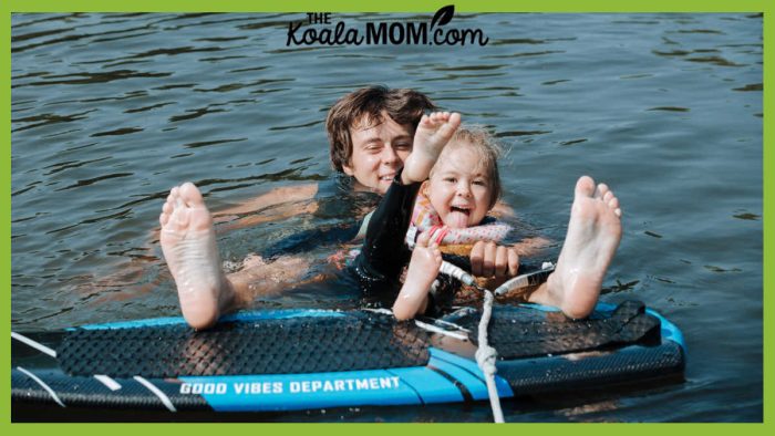 Photo of teenager and younger child playing in lake, with their feet up on a paddleboard, by Ron Lach via Pexels.